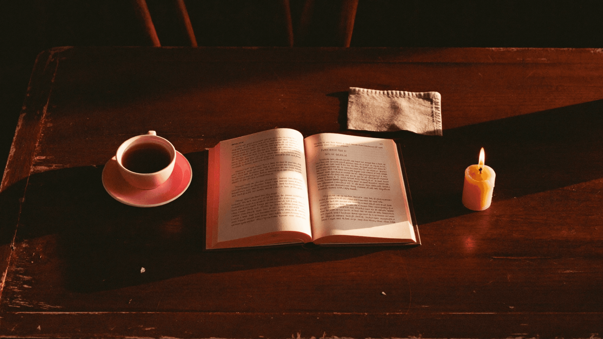 A small lit candle on a wooden cafe table beside an open book and a coffee cup, warm late-afternoon light