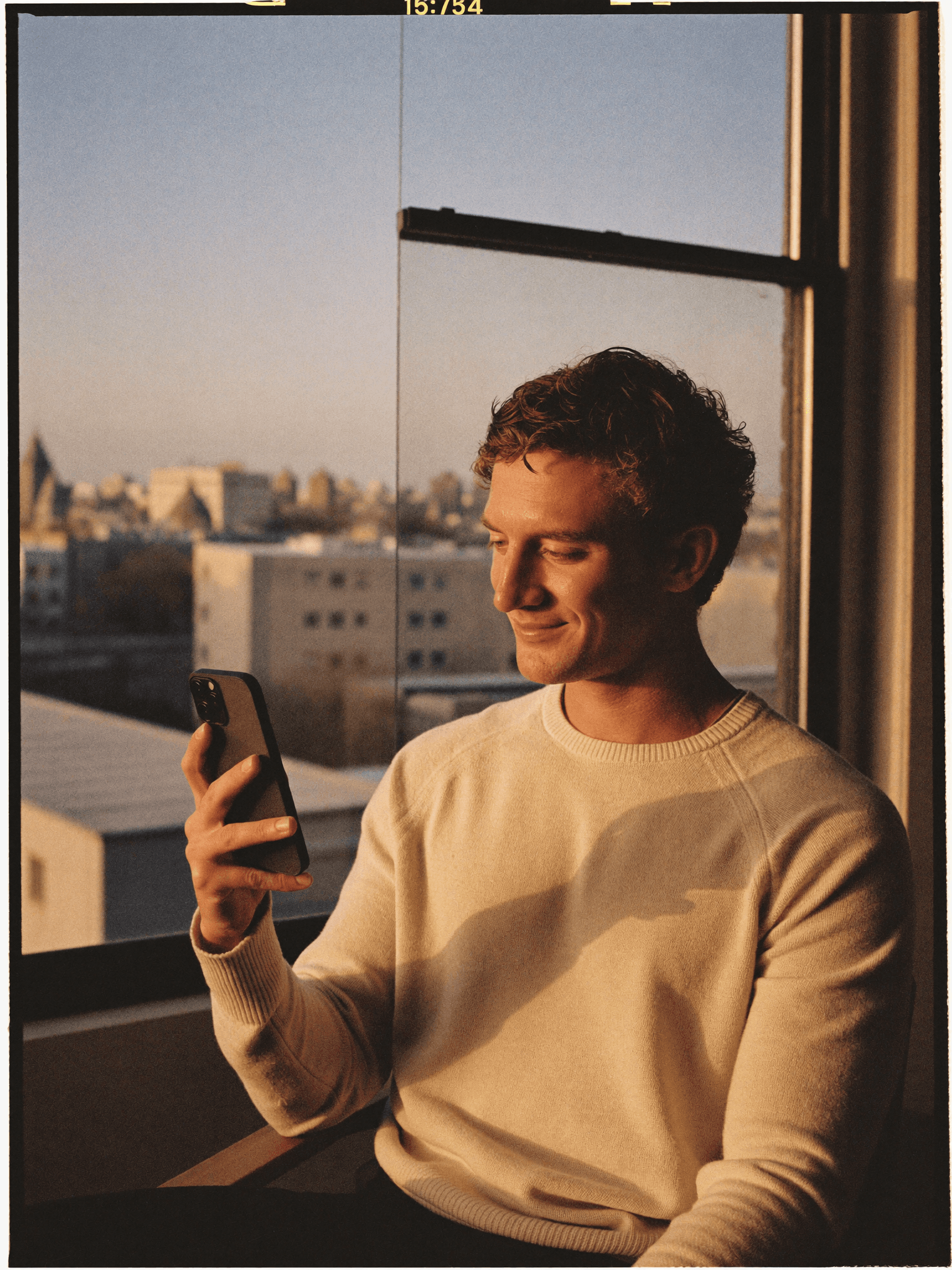 Man by a large window at golden hour holding a phone with a thoughtful smile