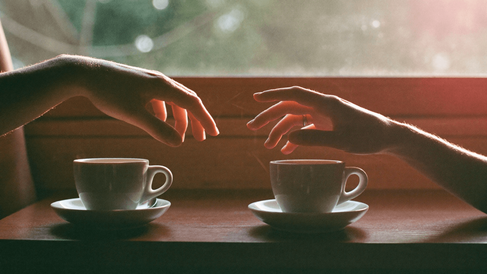 Two hands reaching across a small wooden cafe table with two coffee cups, golden hour light